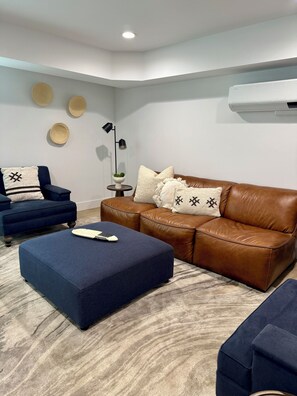 A living room with a blue sofa, a brown leather couch, and a blue ottoman. There is a floor lamp and a small table with a plant. - Beach Haus~Modern Comfort Spacious 4-bedroom in Pacific Beach/La Jolla (San Diego)