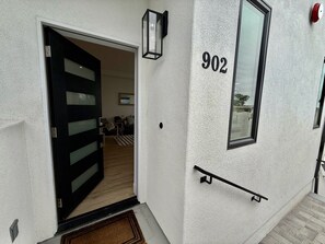 Ein Gebäudeeingang mit einer schwarzen Tür, der Hausnummer '902' an der Wand, einem Fenster und einem roten Feuermelder.