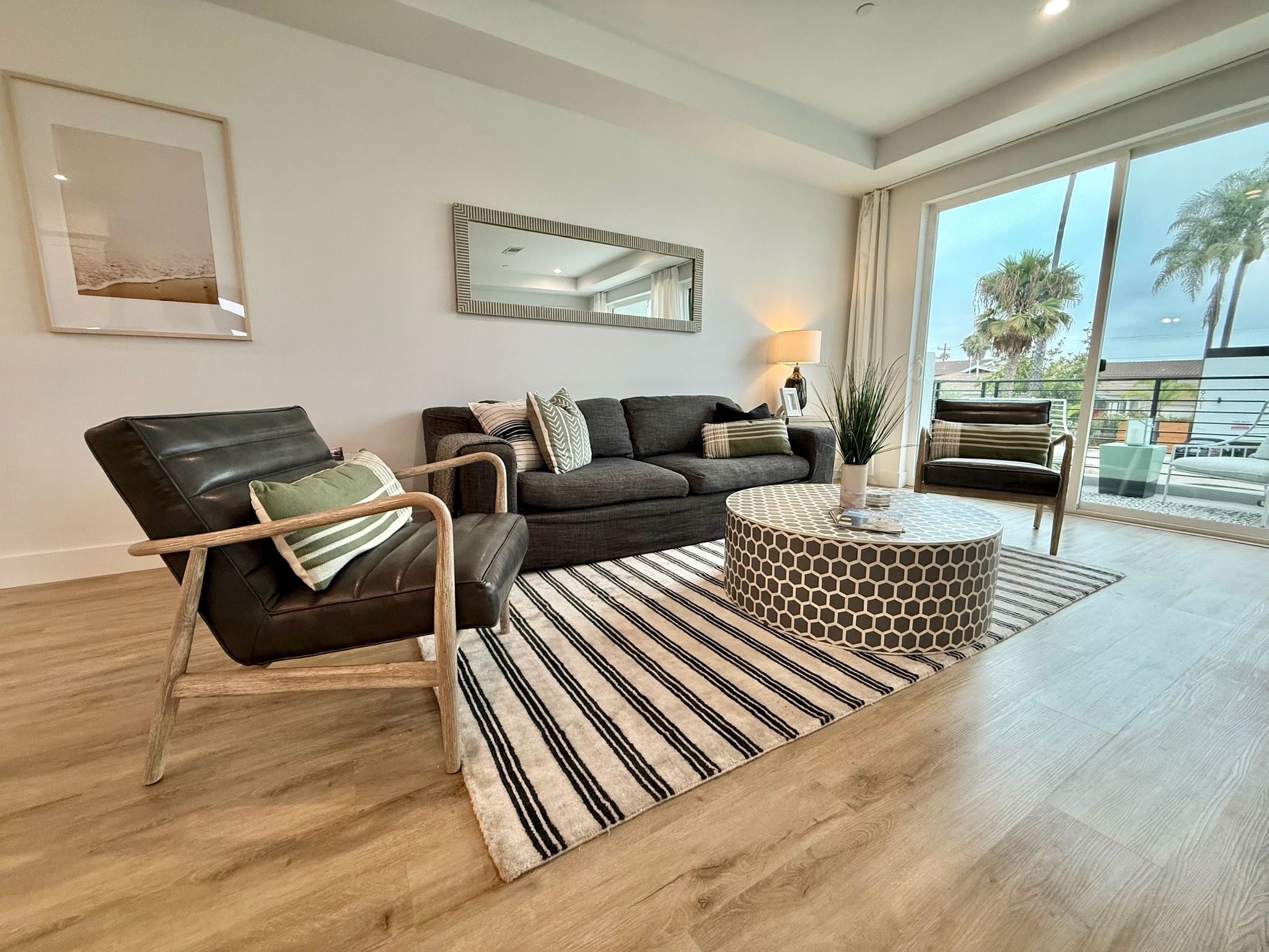 A modern living room with a leather armchair, a dark sofa, a patterned ottoman, and a large window with a view of palm trees.