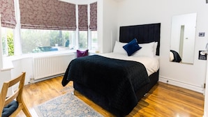 Un dormitorio con una cama grande, una ventana con cortinas, un radiador, suelo de madera y una silla.