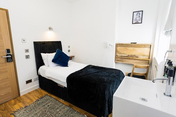 A hotel room with a bed, a bedside table, a desk, a chair, a sink, and a door. - Cambridge House Hotel (Cambridge)