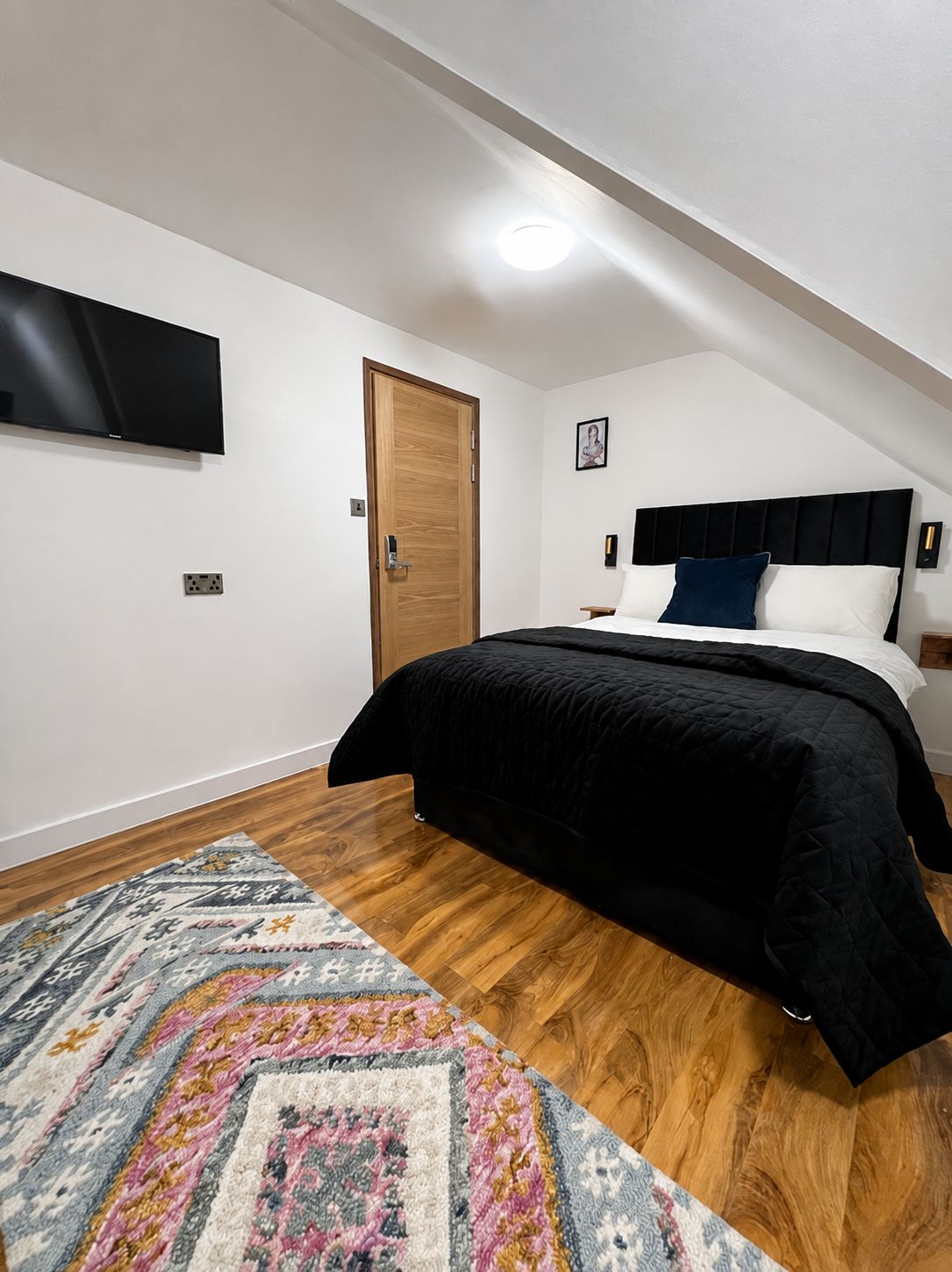 A bedroom with a bed, a flat-screen TV mounted on the wall, a wooden door, and a patterned rug on the floor.