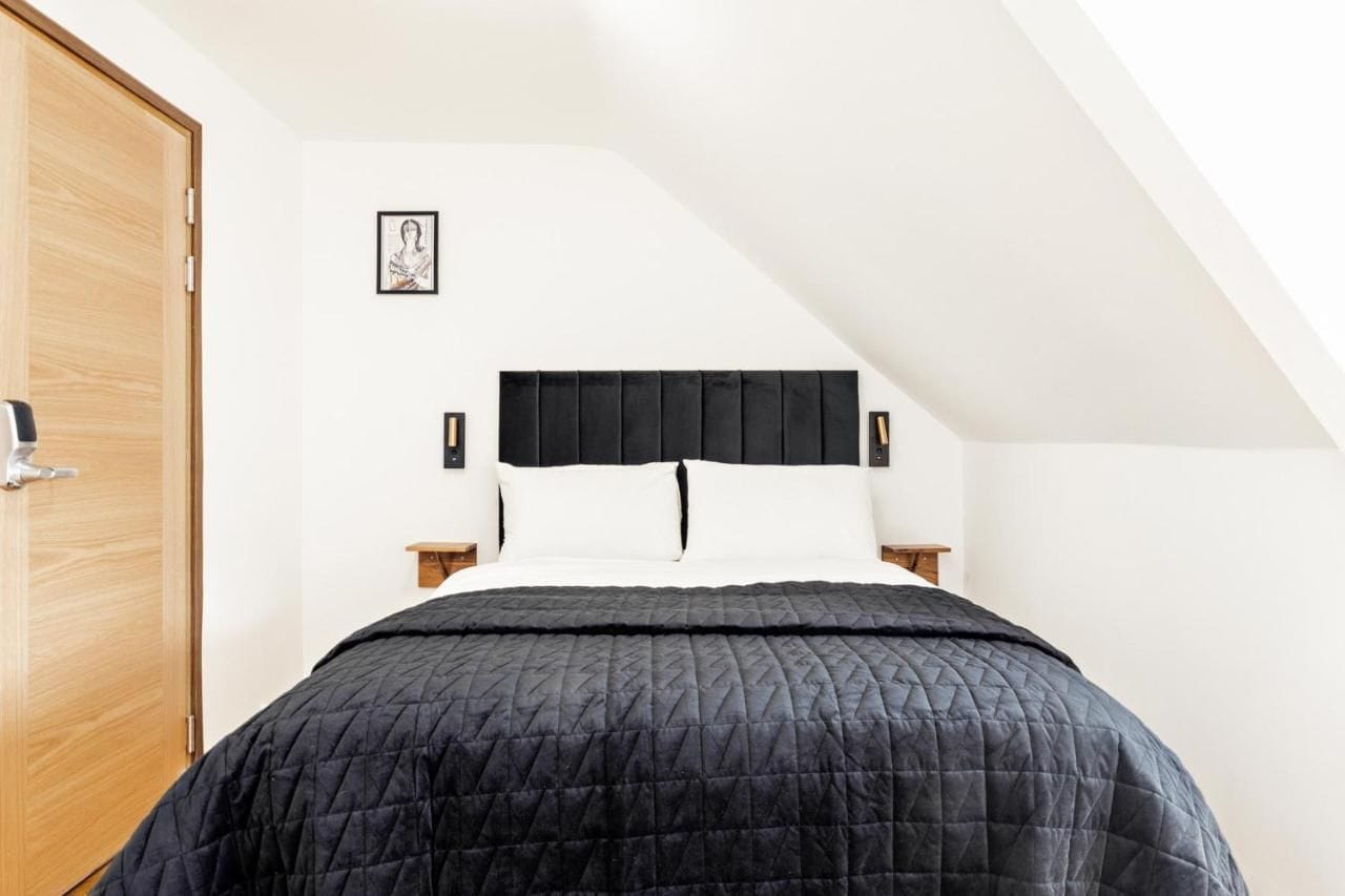 Et soveværelse med skrå vægge, en seng med sort sengegavl, hvide puder og et sort quiltet dynebetræk. En trädør er placeret til venstre, og et lille sengebord med en lampe står ved sengen.