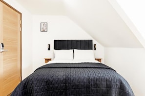 A bedroom with a sloped ceiling, a bed with a black headboard, white pillows, and a black quilted bedspread. There is a wooden door on the left and a small bedside table with a lamp. - Cambridge House Hotel (Cambridge)
