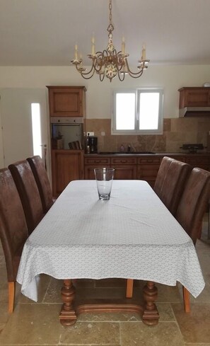 Une table à manger recouverte d’une nappe à motifs, entourée de chaises en bois, dans une cuisine agrémentée d’un lustre et de meubles de rangement en bois.