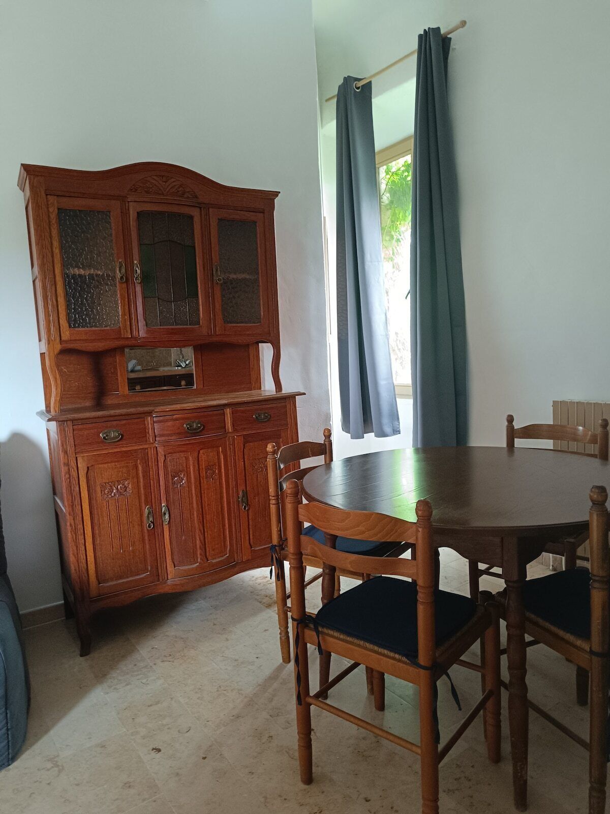 Un comedor con una mesa y sillas de madera, un aparador con puertas de vidrio y una ventana con cortinas.