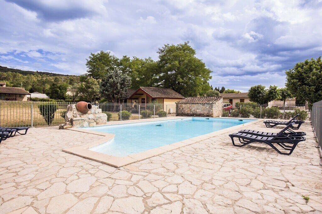Una piscina con baldosas de piedra, sillas de descanso y una zona vallada con un pequeño edificio.