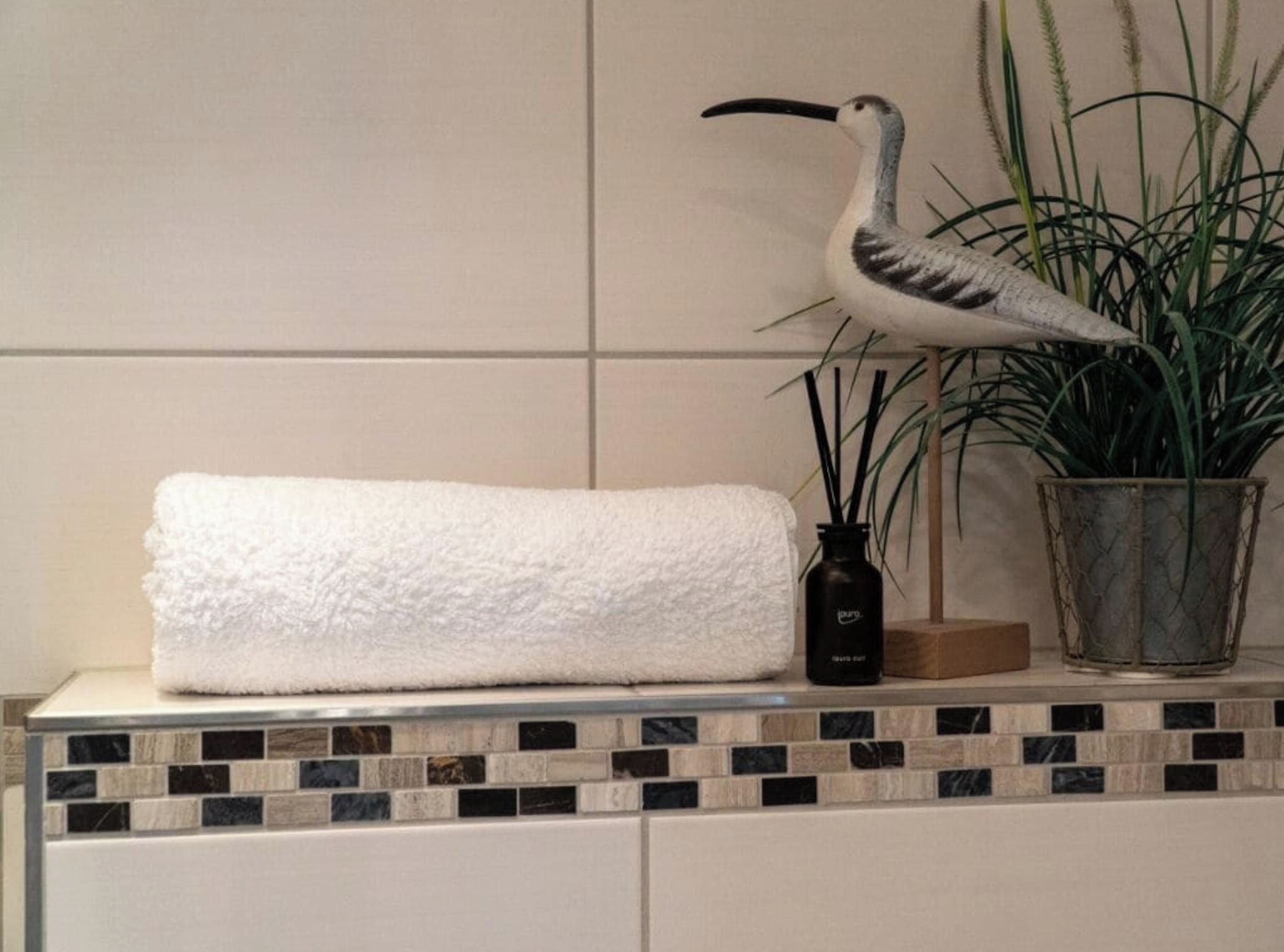 Une salle de bain avec une serviette blanche posée sur un comptoir carrelé, une plante en pot et une bouteille noire.
