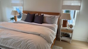 A neatly made bed with a wooden headboard, two bedside tables with lamps, and a window with a view of buildings. - Summersea 3-bedroom house on the water in soothing New Seabury, Mashpee (Mashpee)