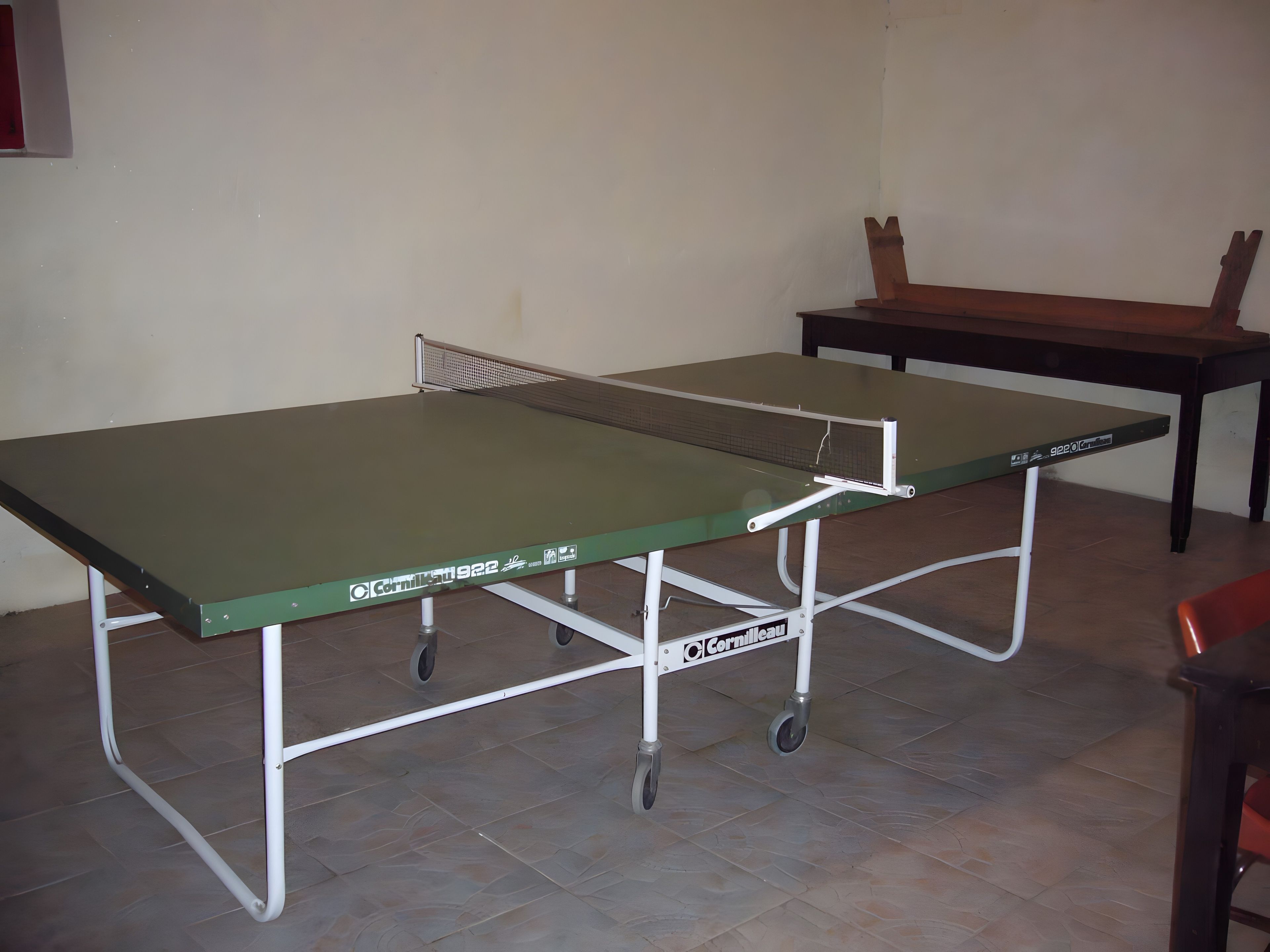 A table tennis table with wheels, a chair, and a wooden table in a room with tiled flooring.