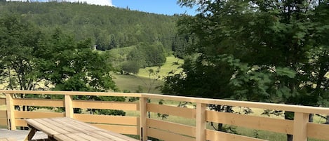 Una mesa de picnic de madera con bancos, sobre un lecho de grava, con una baranda de madera al fondo y una ladera boscosa en la distancia.