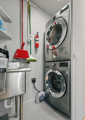 A laundry room with two stacked front-loading washing machines, a red mop, a green broom, and a fire extinguisher on the wall. - Blazing Elephant - Stunning VIEWS, Pet Friendly, Central Location (Sedona)