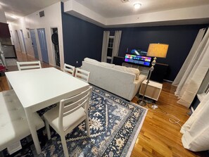 A modern living room with a white sofa, a dining table with chairs, a patterned rug, and a flat-screen TV. - 3-bed/2-bath apt in Jersey City mins from NYC/American Dream/Jets & Giants Arena (Jersey City)