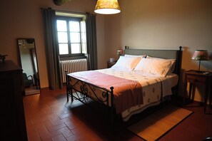 Ein Schlafzimmer mit einem Bett, einem Heizkörper, einem Fenster mit Vorhängen, einer Nachttischlampe, einem Spiegel und einem Holzboden.