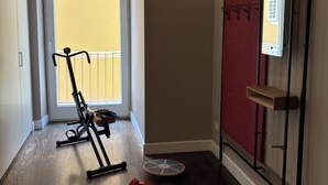 Huoneessa on puulattia, ovi, joka johtaa parvekkeelle, pystyssä oleva kuntopyörä, pari käsipainoja ja seinälle kiinnitetty hylly koukuilla.