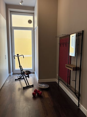 A room with a wooden floor, a door leading to a balcony, a standing exercise bike, a pair of dumbbells, and a wall-mounted rack with hooks. - Vrbo Property (ITALIA)