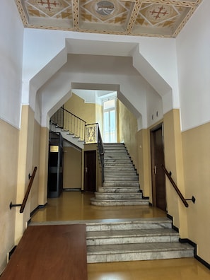 Un grand escalier avec des marches en marbre et des rampes noires, menant à un palier doté d’une porte et d’une fenêtre.