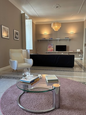 A modern living room with a glass coffee table, a white armchair, a black sofa, and a television on a wall-mounted shelf. - Rattazzi 36 - La Spezia & 5 Terre (ITALIA)