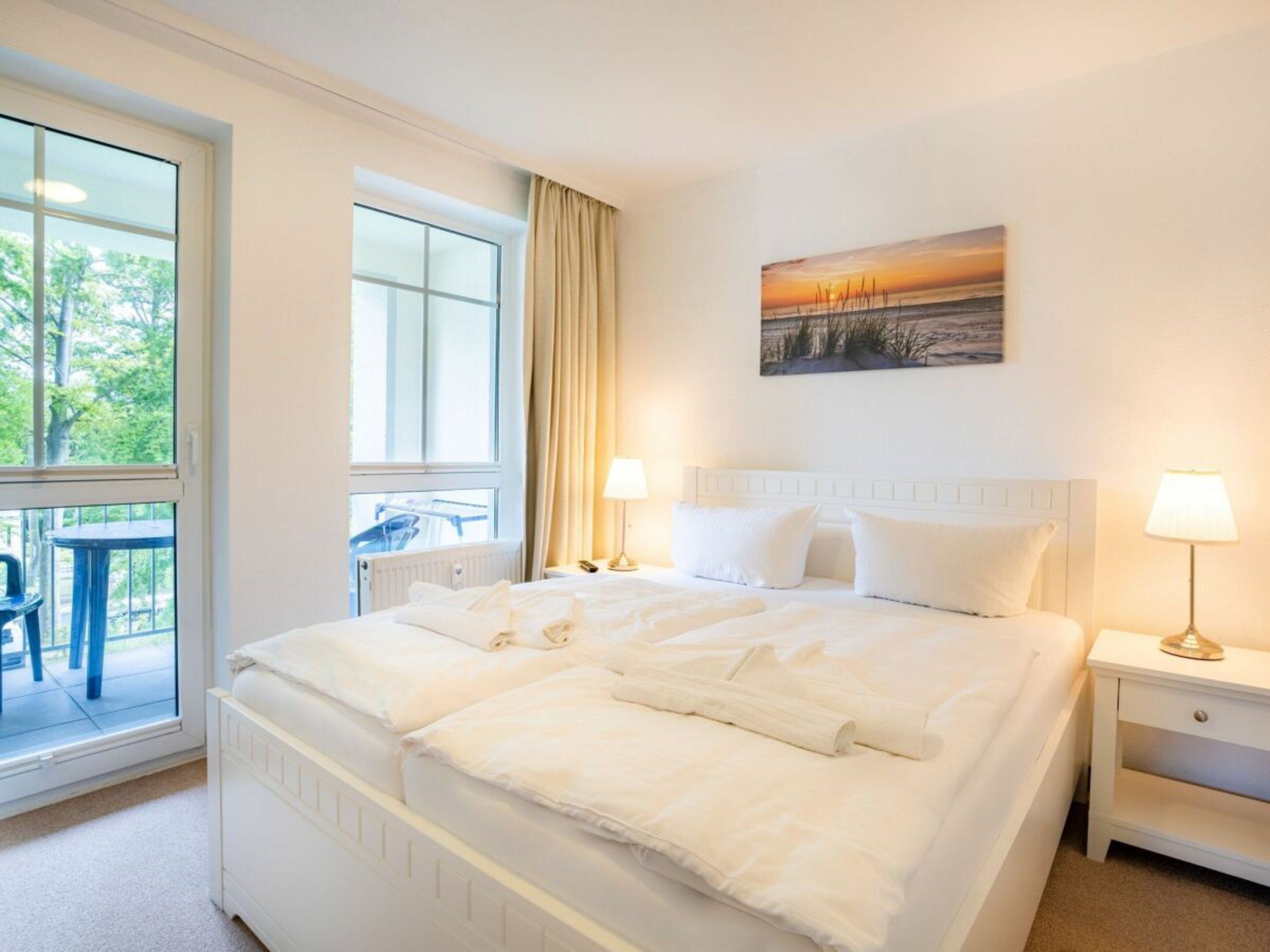 A hotel room with a large bed, two bedside tables with lamps, a window with a view of greenery, and a painting on the wall.