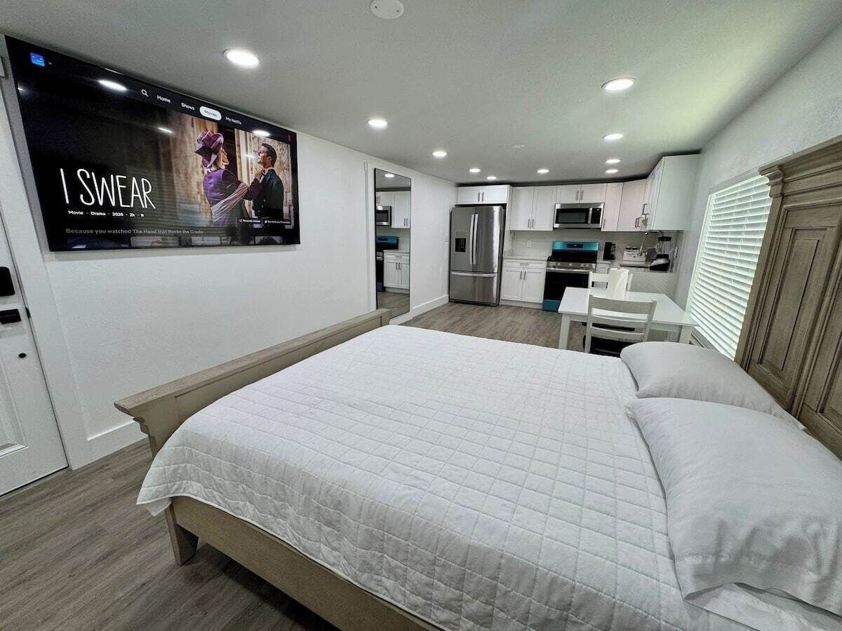 A modern bedroom with a large bed, a kitchenette, and a TV mounted on the wall.