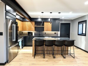 A modern kitchen with a stainless steel refrigerator, wooden cabinets, a black backsplash, and a bar area with stools. - Upscale Loft (Thunder Bay)