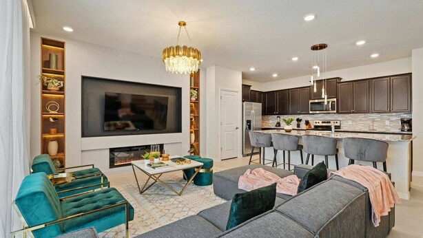 A modern living room with a large flat-screen TV, a fireplace, teal and gray sofas, and a kitchen area with dark cabinets and a stainless steel refrigerator.