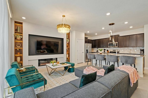 A modern living room with a large flat-screen TV, a fireplace, teal and gray sofas, and a kitchen area with dark cabinets and a stainless steel refrigerator.
