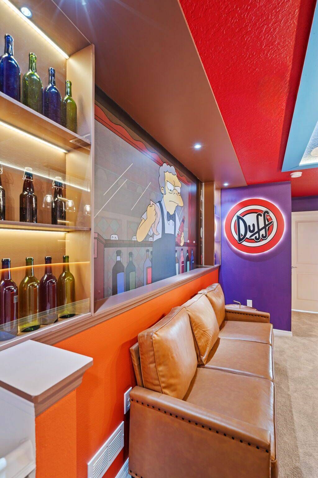 Uma área de bar com sofá de couro, parede com personagem de desenho animado e placa 'Duff'.