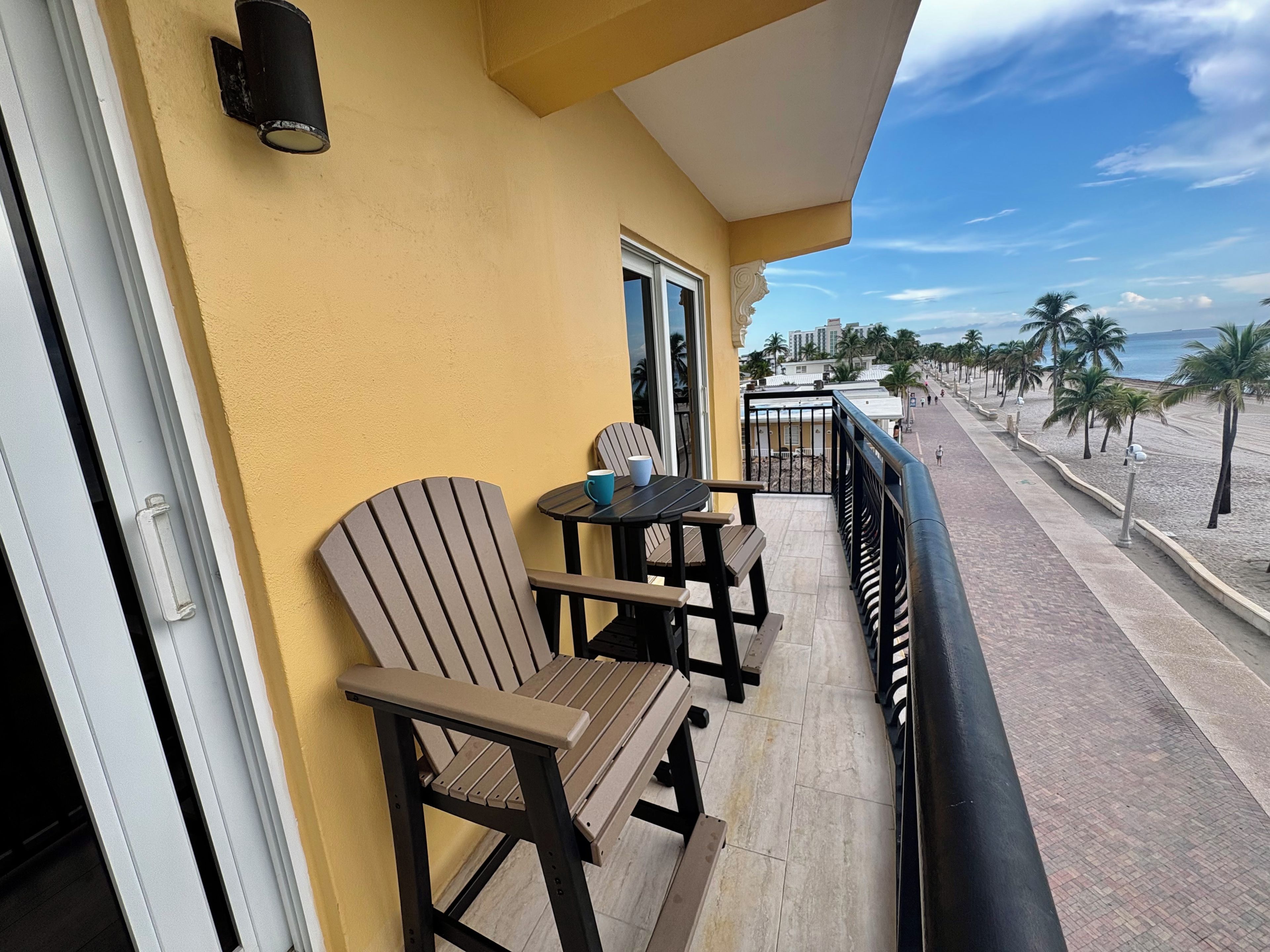 A balcony with two chairs and a table, overlooking a beach with palm trees and a clear sky.