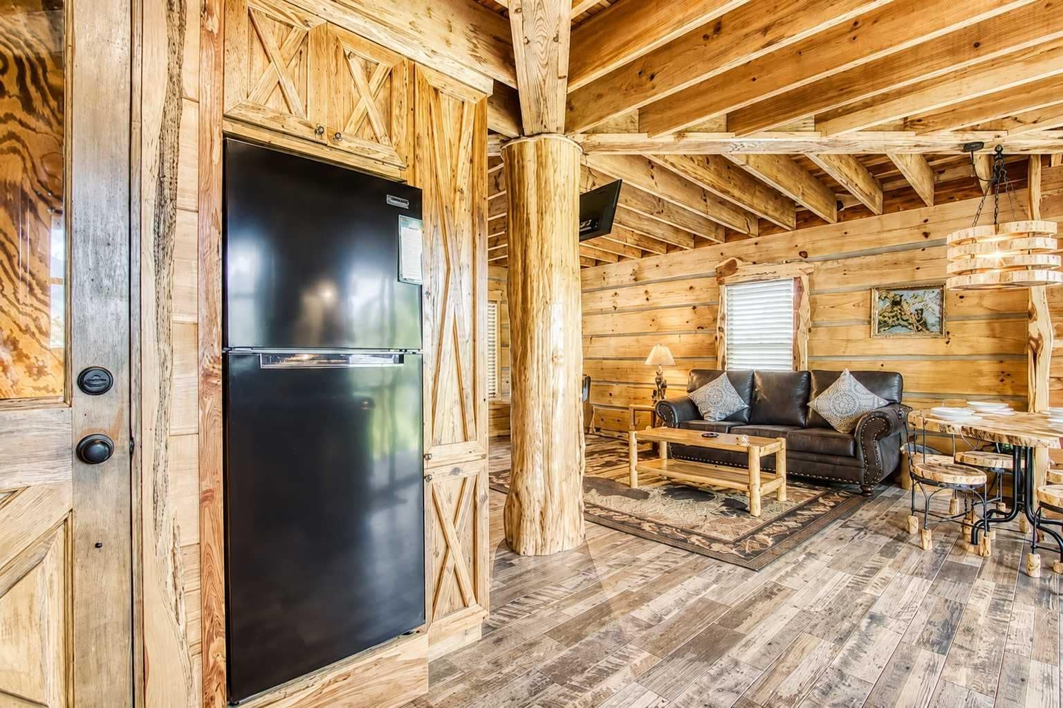 Interior de una cabaña de madera con un refrigerador negro, un sofá y una mesa de comedor.