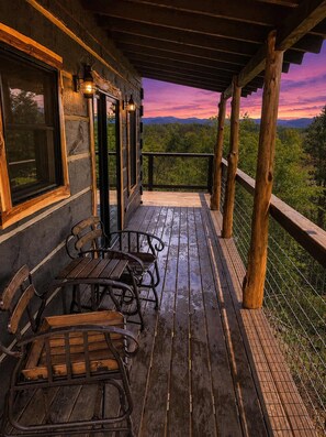 A wooden deck with a view of a forested landscape, a covered seating area with metal chairs, and a railing. - 2-Bedroom Castle Cabin with Dungeon Game Room, Hot Tub & Mountain Views (Sevierville)