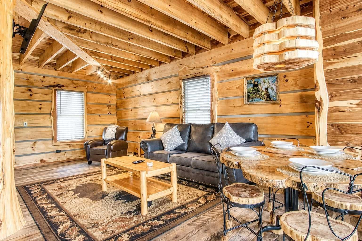 Ein Holzblockhaus-Innenraum mit einer Couch, Sesseln, einem Couchtisch, einem Esstisch mit Stühlen und einer Hängelampe.