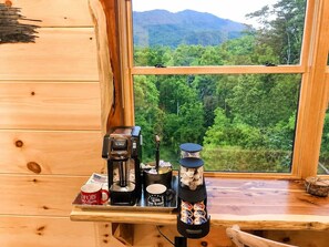 Ein Interieur einer Holzhütte mit Kaffeemaschine, Tassen und einer Snacksammlung auf einem Holztisch neben einem Fenster mit Blick auf einen Wald.