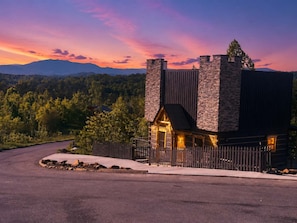 A modern building with a stone facade and a wooden entrance, surrounded by a fence and a road, with a mountainous landscape in the background. - 2-Bedroom Castle Cabin with Dungeon Game Room, Hot Tub & Mountain Views (Sevierville)