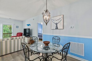 A dining area with a glass table and metal chairs, a red sofa, a television, and artwork on the walls. - Whale of a Tale | Beach Getaway + Pool + Screen (North Myrtle Beach)