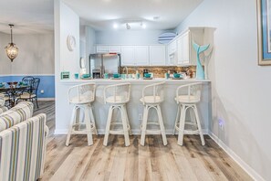 A kitchen with white cabinets, a central island with bar stools, and a blue decorative piece on the wall. - Whale of a Tale | Beach Getaway + Pool + Screen (North Myrtle Beach)