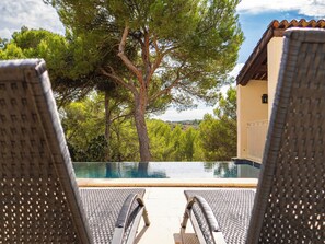 A poolside view with two lounge chairs facing a swimming pool, surrounded by trees and a building with a tiled roof. - Le Petit Mas - Piscine - Golf de Pont Royal (Mallemort)