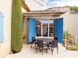 En udendørs terrasse med et rundt bord og stole, en pergola og udsigt til gaden.