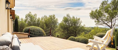 A deck with wicker furniture and a chair, overlooking a landscaped garden with trees and a mountain in the distance.