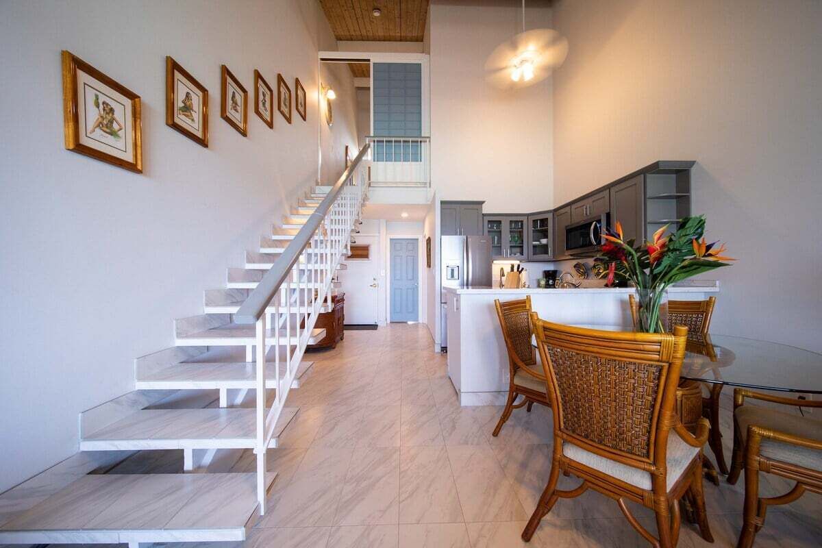 A modern kitchen with white cabinets, a dining area with wicker chairs, and a staircase leading to an upper level.