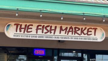A storefront with a sign reading "The Fish Market" offering seafood, marinades, salads, fish burgers, and cafe dining in air-conditioned comfort.