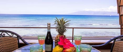 A table with a bottle of Jamaica wine, pineapple, mango, and a red flower, with two glasses of red drink, overlooking the ocean.