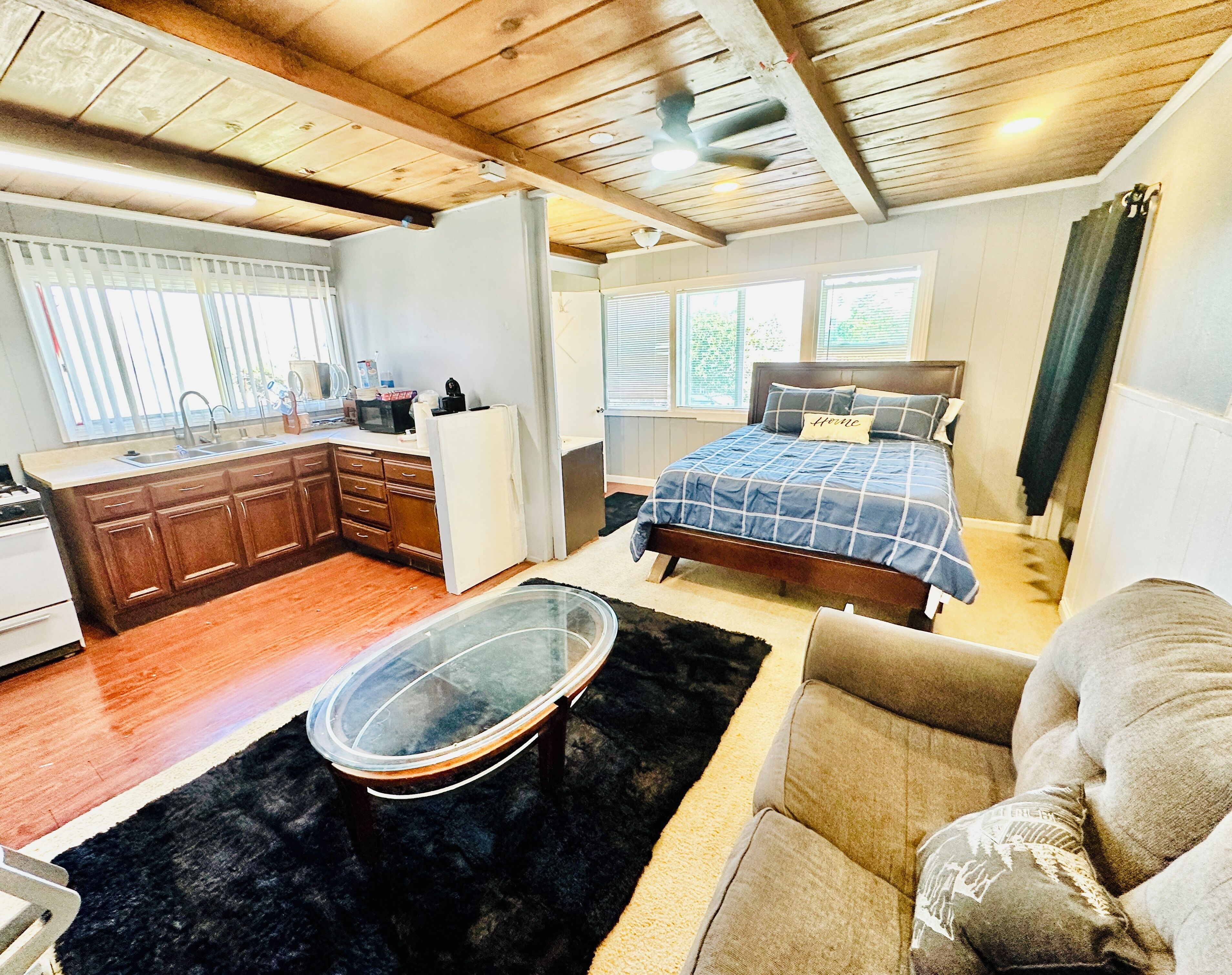 A compact room with a kitchenette, a bed with a blue checkered bedspread, and a small seating area with a glass-top table.