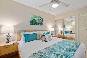 Ein ordentlich eingerichtetes Schlafzimmer mit einem Bett, zwei Nachttischen, einem Deckenventilator, einem großen Spiegel und einem an der Wand hängenden Kunstwerk.
