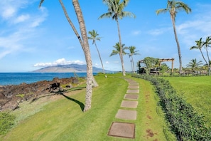 A well-maintained grassy area with a stone pathway leading to a wooden pergola. - Updated Beachfront 1BD w Ocean Views - Kihei Surfside 306 (Kihei)