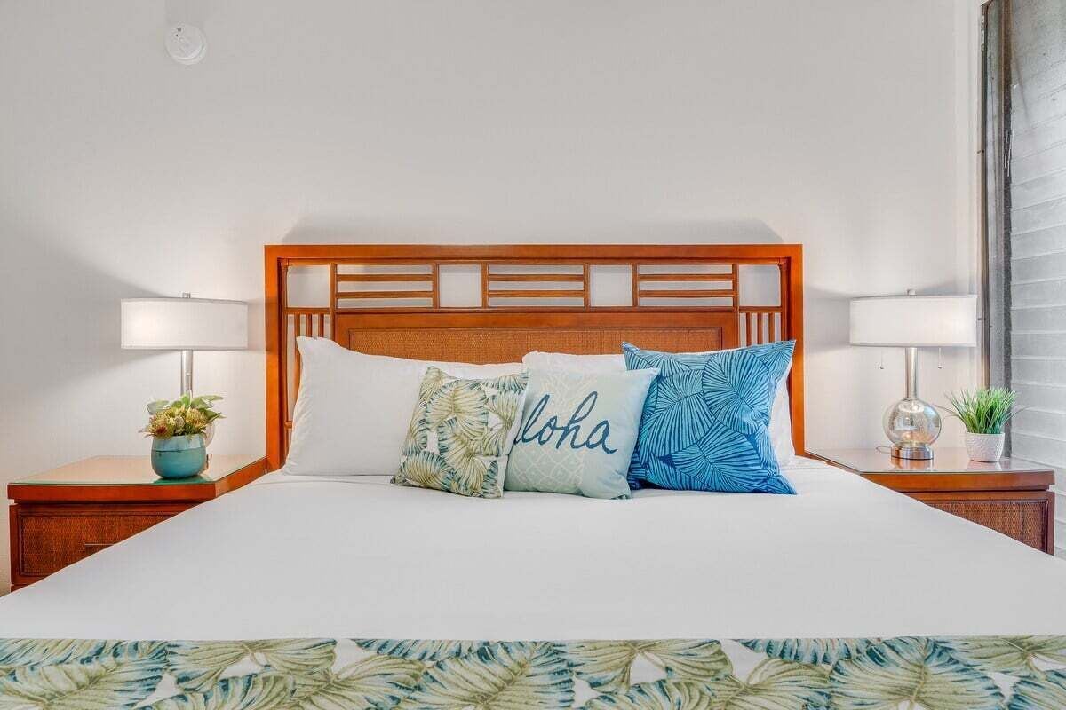 A neatly made bed with a wooden headboard, multiple pillows, and bedside tables with lamps and small plants.