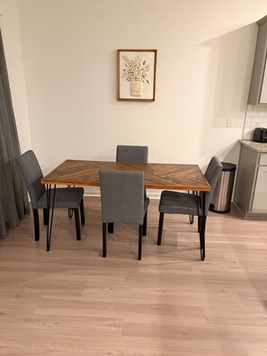 Zona pranzo con tavolo in legno e sei sedie, un quadro appeso al muro e un angolo cottura sulla destra.