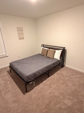 Ein Einzelbett mit Metallgestell und grauer Bettwäsche in einem Zimmer mit beigem Teppichboden und weißen Wänden.