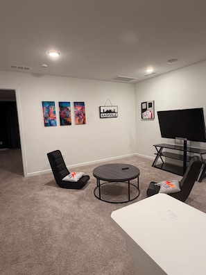 Ein Wohnzimmer mit Flachbildfernseher, einem runden Couchtisch, zwei schwarzen Sesseln und einem Tisch in Weiß. An der Wand hängen drei gerahmte Kunstwerke.