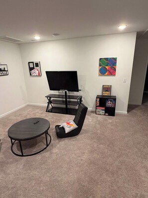 A room with a flat-screen TV on a stand, a black gaming chair, a small table, and a Monopoly game box. - Beautiful Modern Home Near I-65 - Near Nashville (White House)